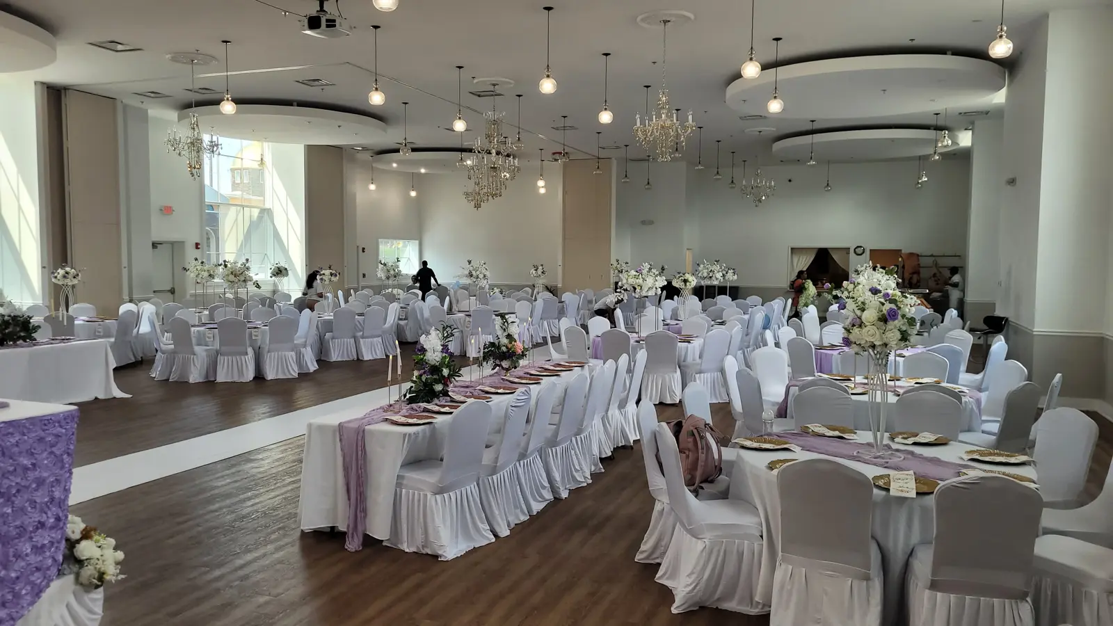 Hall set for a reception with white chair covers and lavender accents under crystal chandeliers